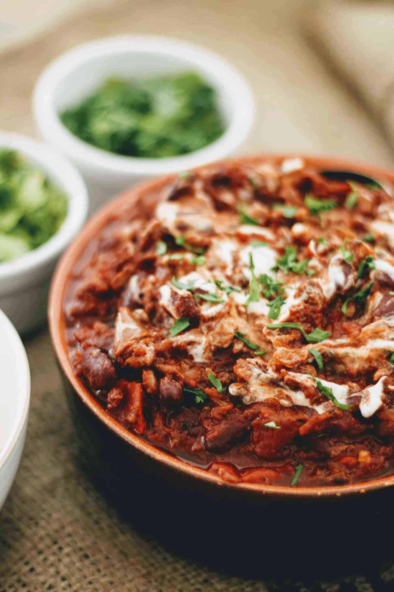 close up of chilli beef in a round terracotta cooking pot with side garnishes of fresh herbs