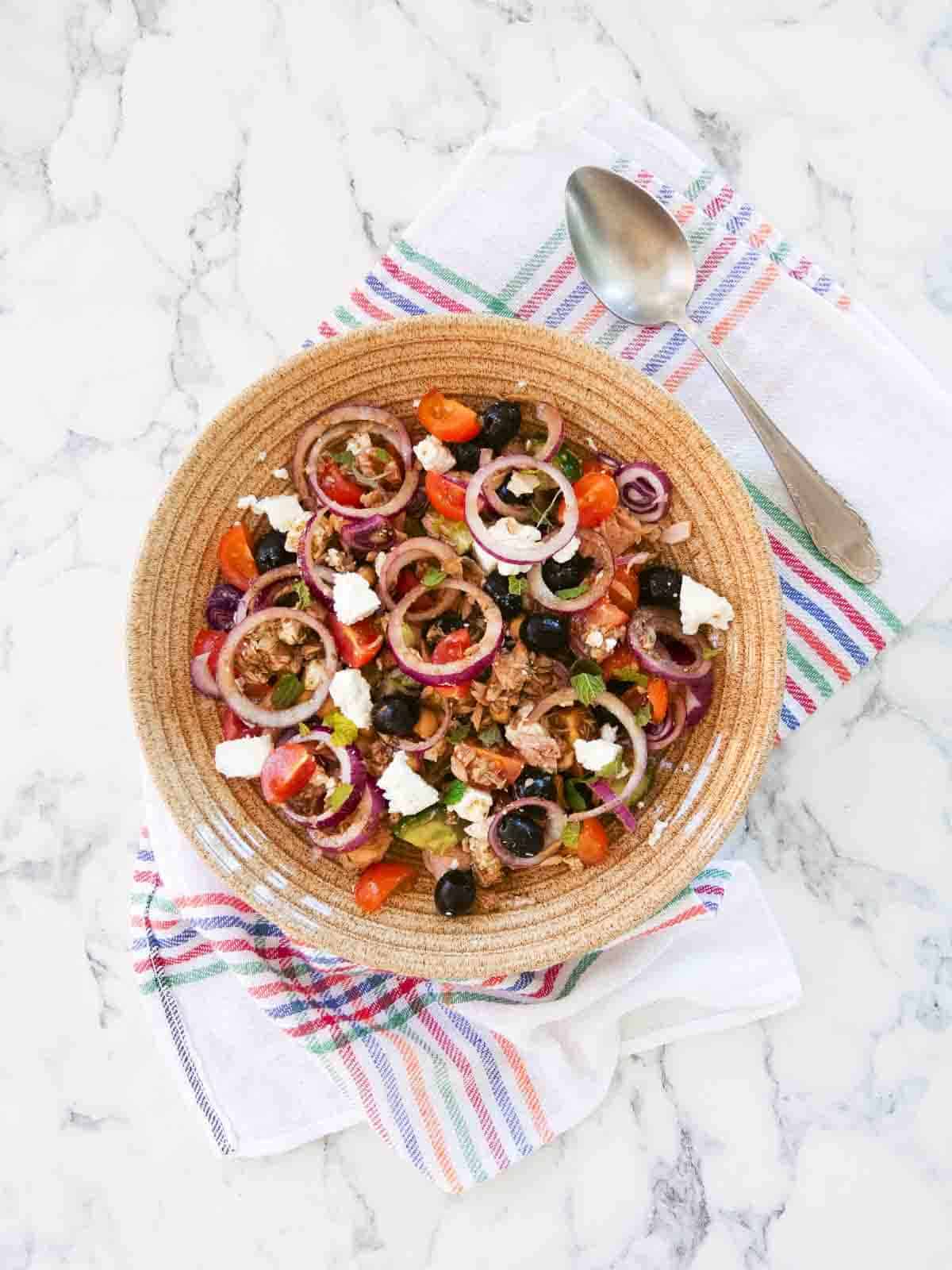 Bowl of salad placed on a large Spanish tea towel in the centre of a marble worktop with spoon, ready for serving