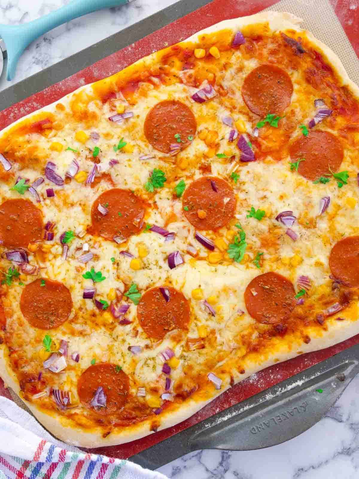 Birdseye view of a thin crust pizza dough recipe shaped in to a square and topped with pepperoni, onions, sweetcorn and fresh herbs