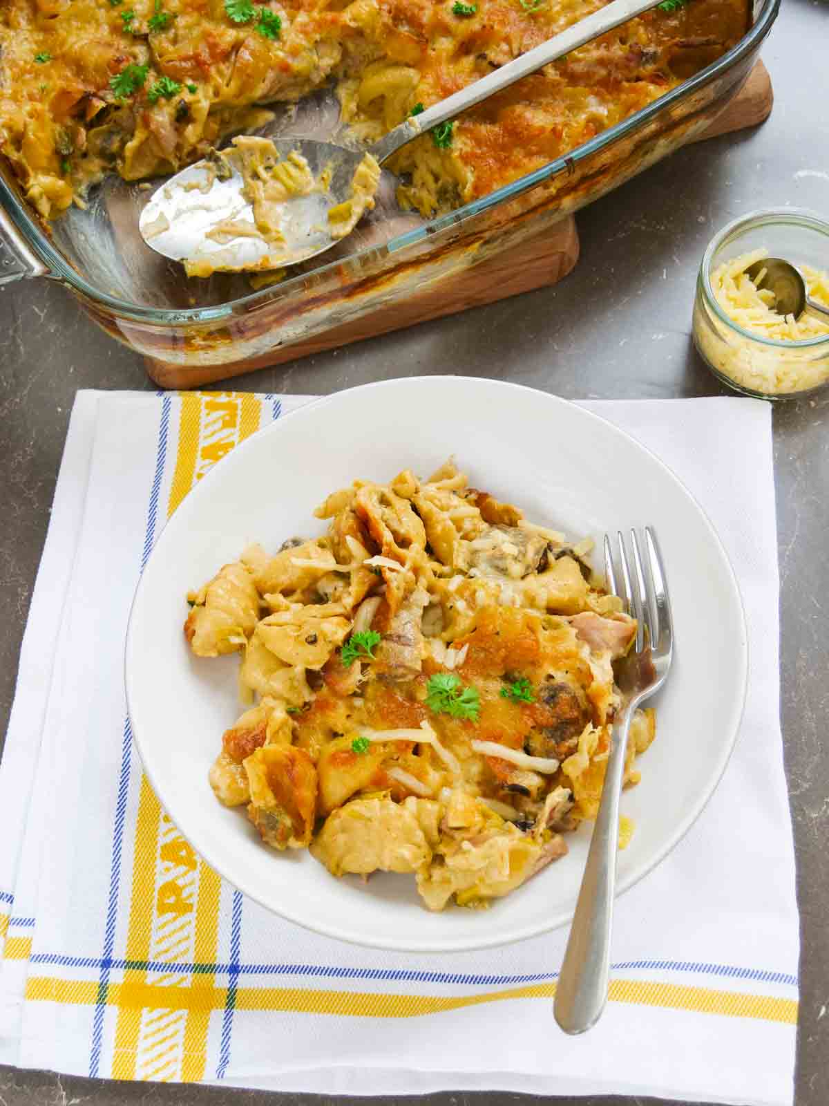 White Denby bowl full of a creamy cheese pasta bake with bacon, leeks and mushrooms, sat on a Spanish tea towel.