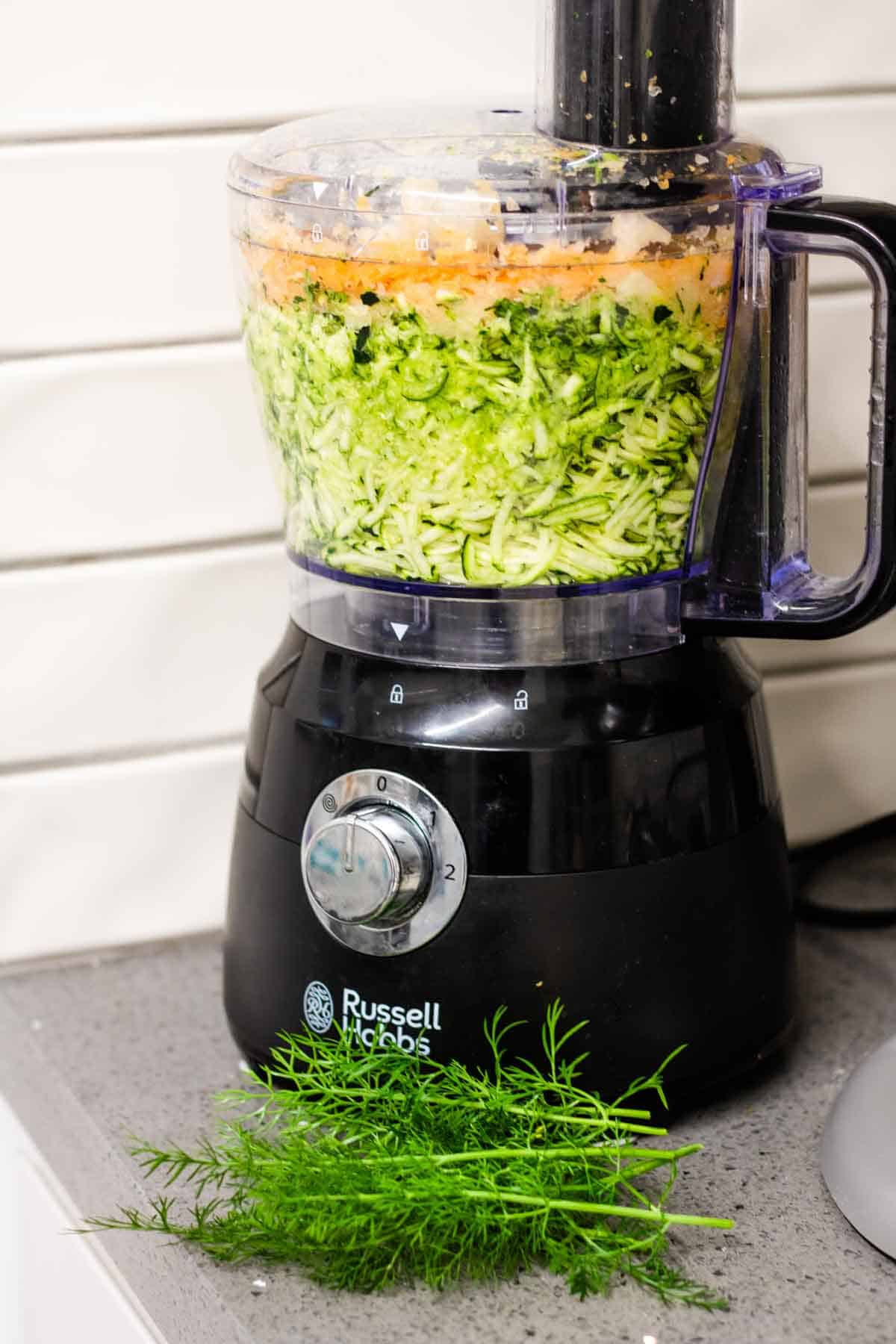 A food processor full of courgettes, carrots and onion to make a shrimp fritters recipe with fennel fronds