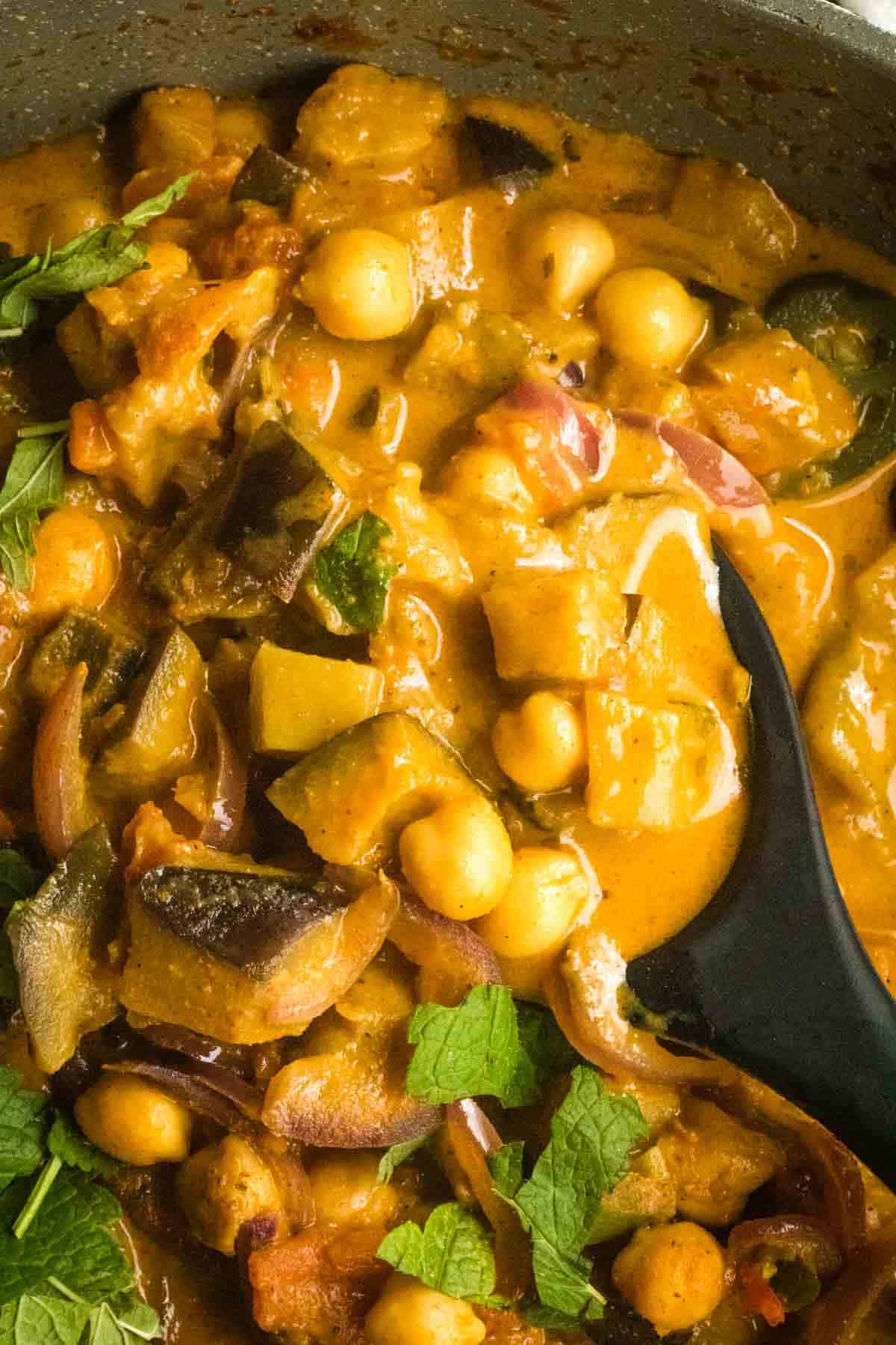 Close up of a black spoon in a pan with a curry aubergine recipe and chickpeas