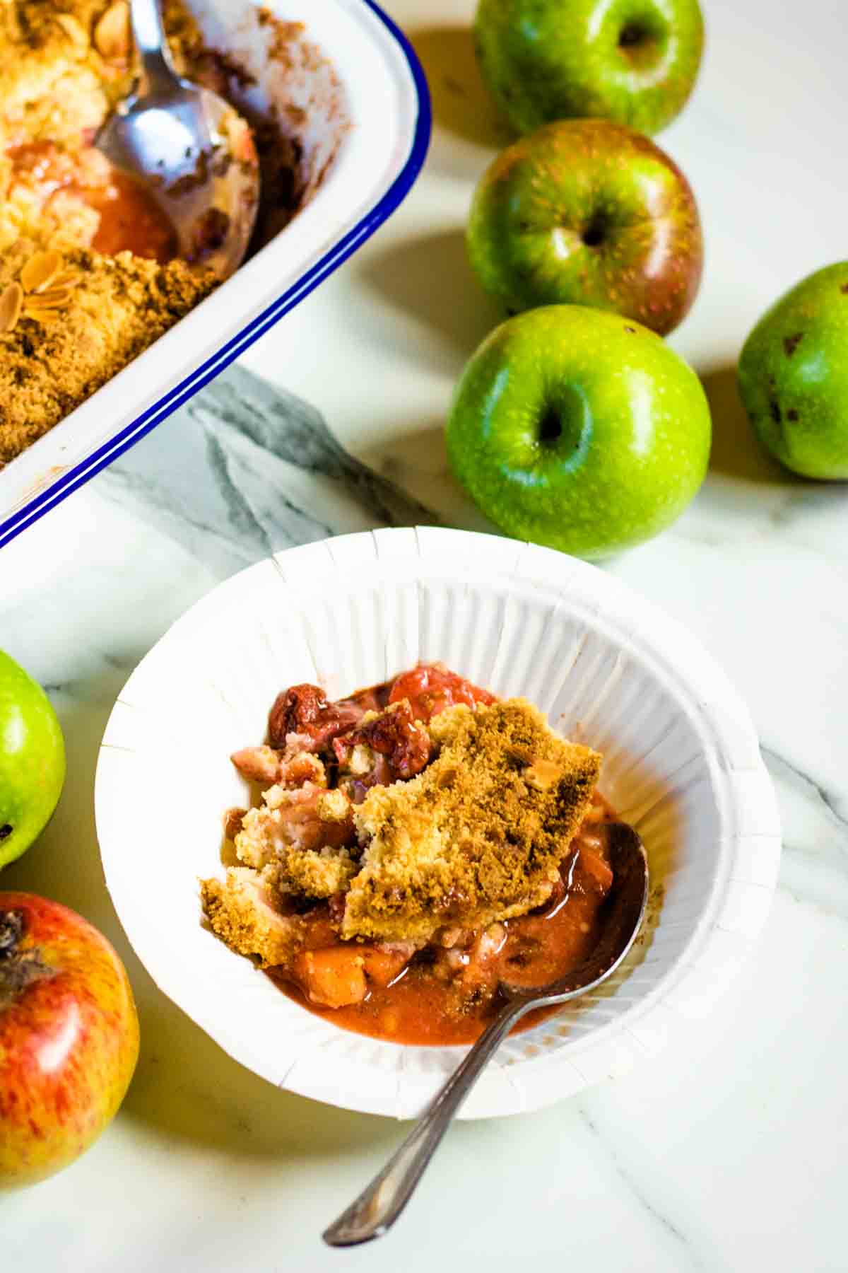 White paper bowl for a party serving of strawberry crunch crumble surrounded by apples