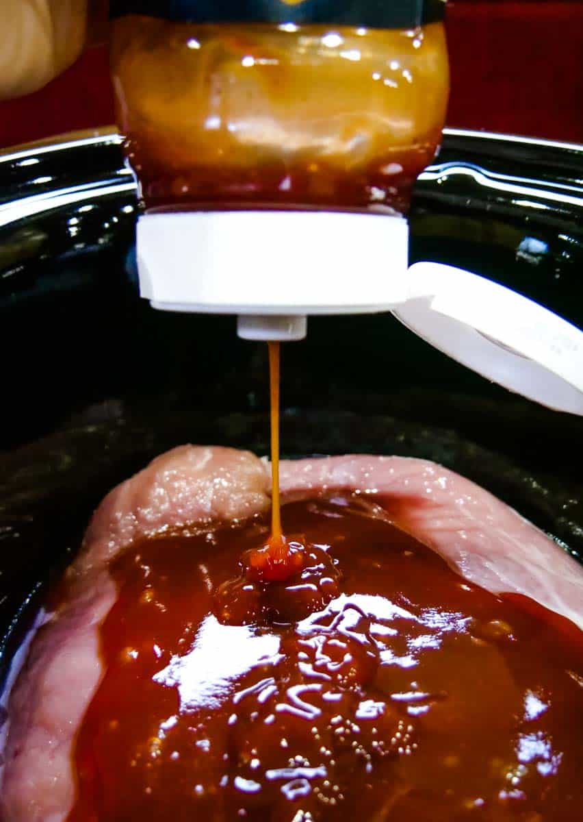 BBQ sauce being poured on top of pork in a slow cooker