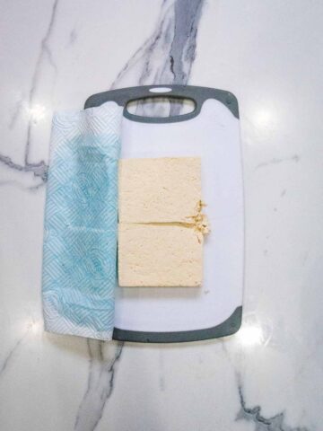 Tofu laid on a chopping board with wet kitchen towel peeled back after pressing