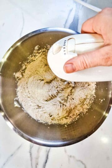 person creaming butter andlight brown soft sugar with a hand mixer in a large stainless steel mixing bowl