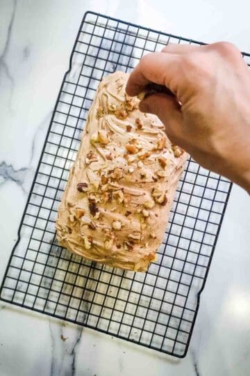 person sprinkling walnut pieces onto a coffee loaf cake with buttercream on top