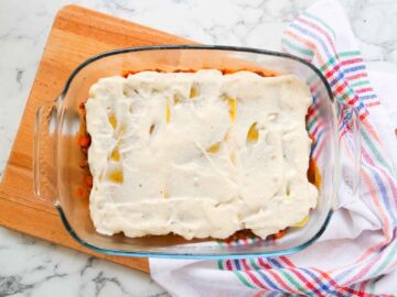 cheesy bechamel sauce spread on top of pasta sheets and beef ragu in glass baking dish