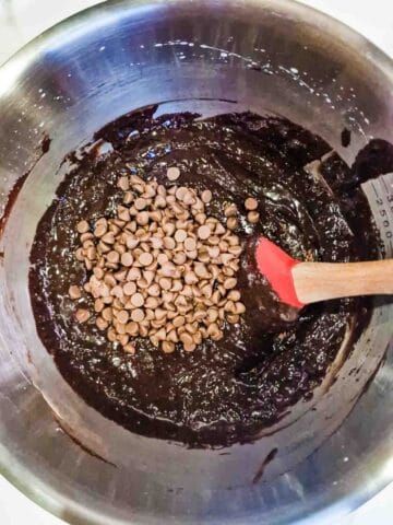 person slowly folding milk chocolate chips in chocolate brownie batter with a spatula in a stainless steel bowl