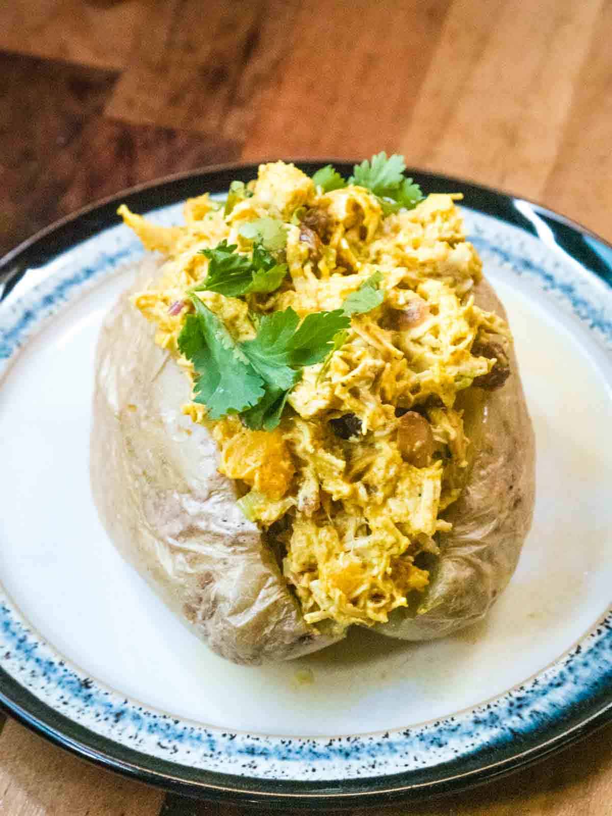 coronation chicken jacket potato on a white and black rimmed plate topped with coriander leaves on a wooden kitchen worktop