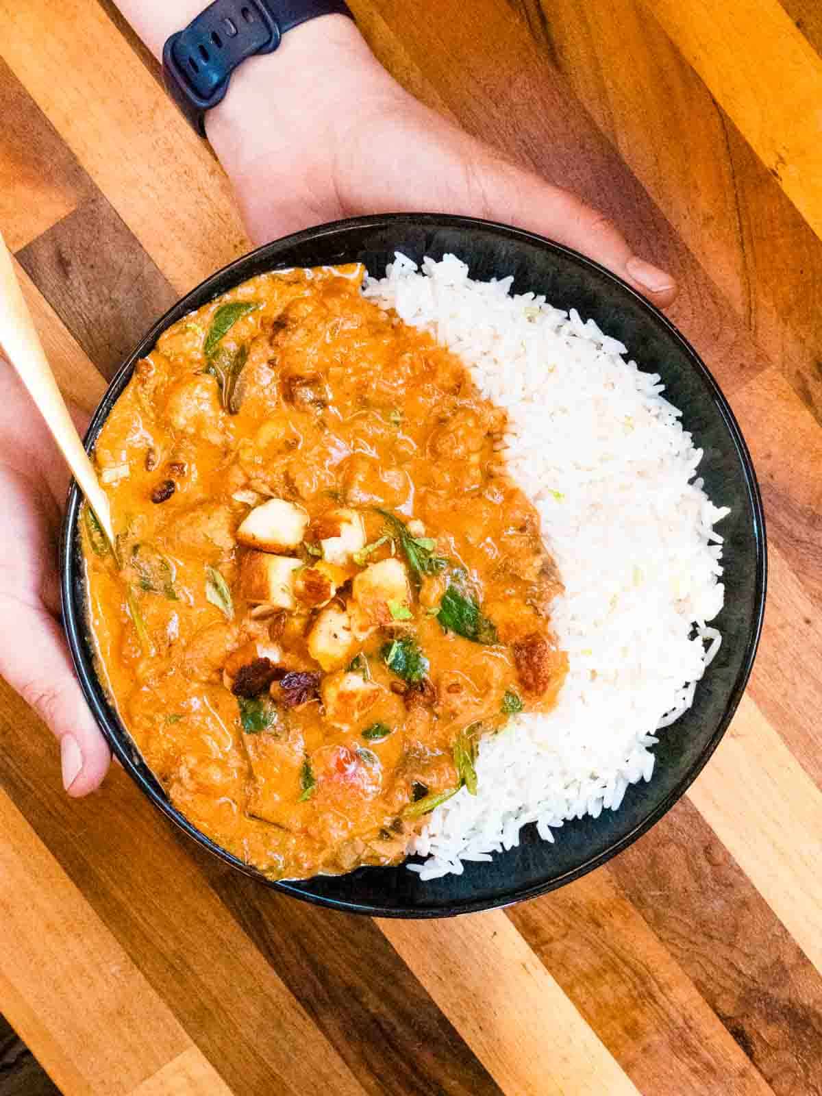 sausage meat curry in a black bowl with white rice in a bowl being held
