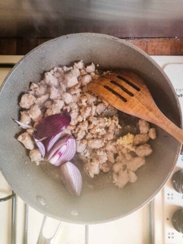 person frying sausage meat in a large frying pan while breaking it up with a wooden spoon and frying onions and garlic