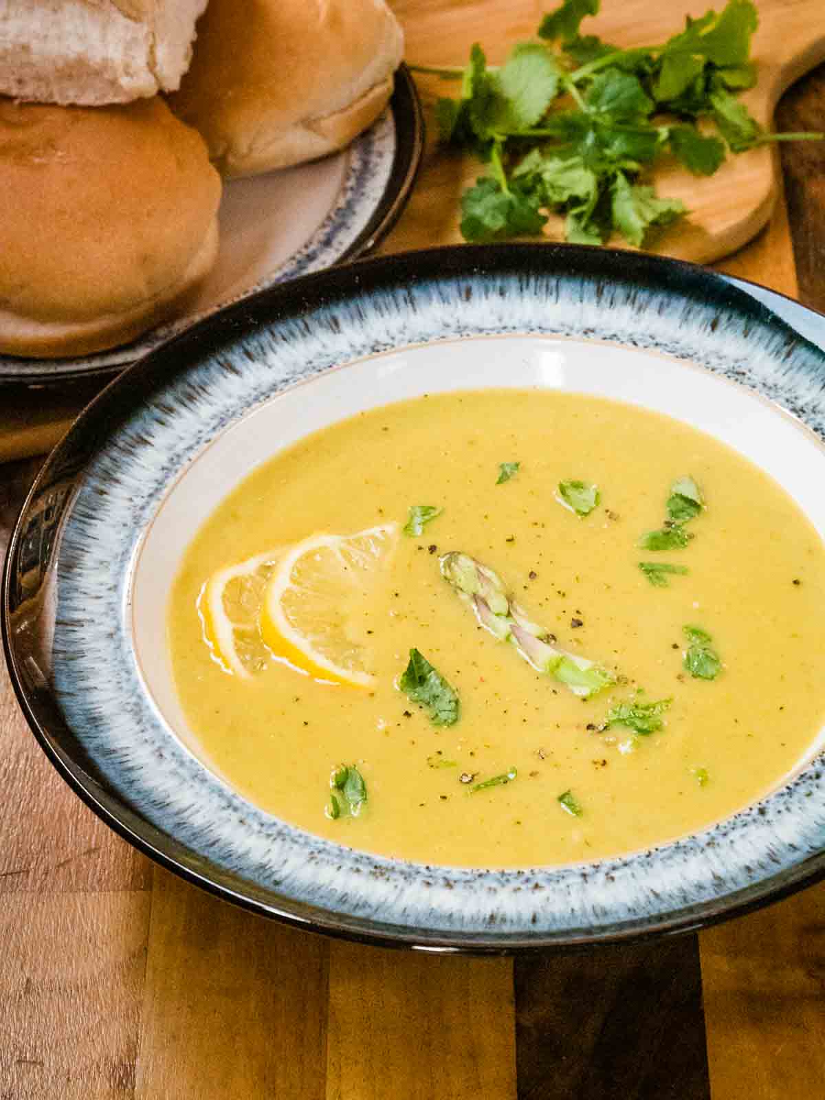 close up of vegan creamy asparagus soup with lemon slices, coriander and seasoning in a large black and white Denby halo bowl on wooden counter