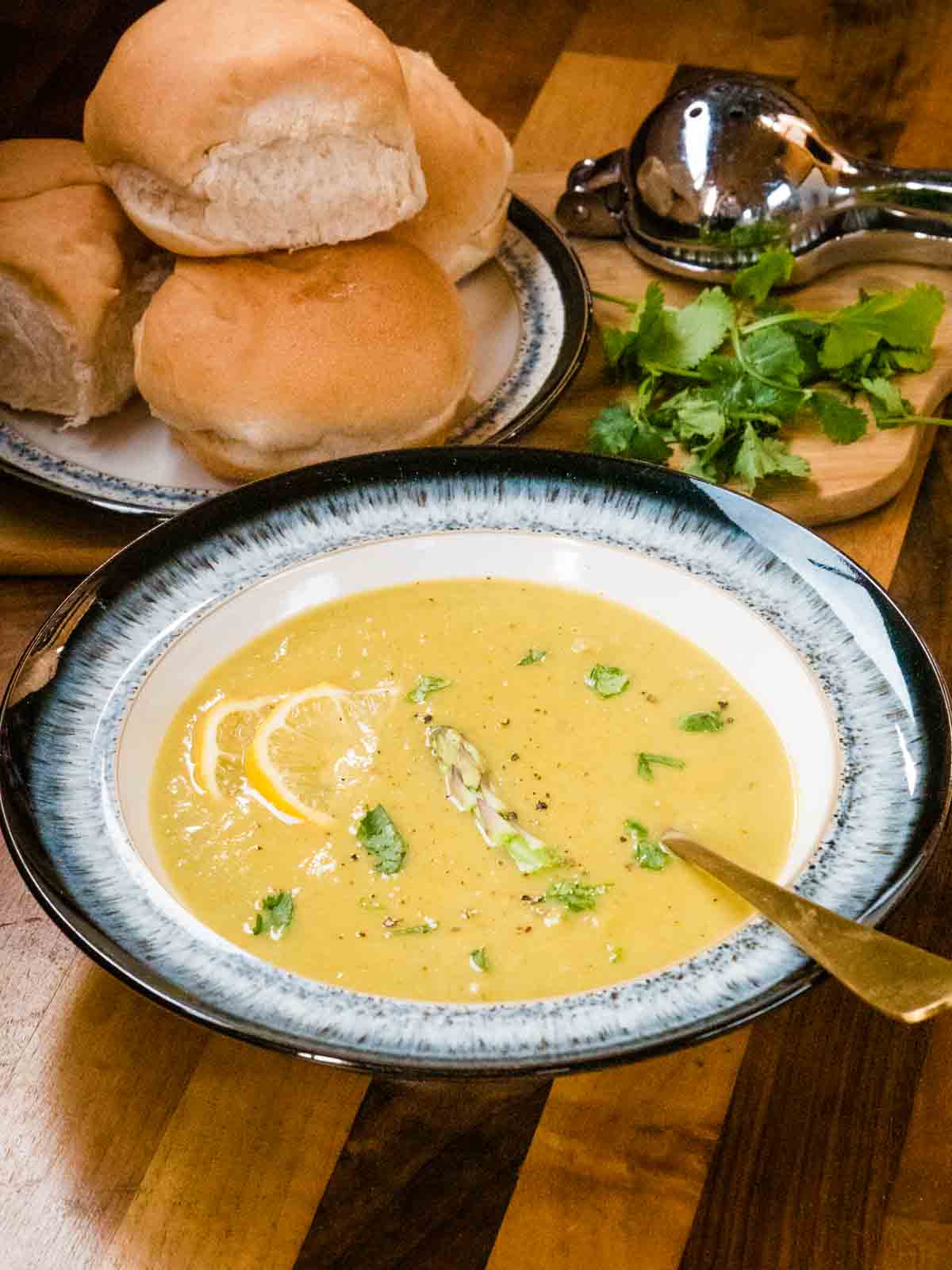 asparagus soup in a bowl surrounded by bread rolls on a plate wit fresh coriander and a lemon juice