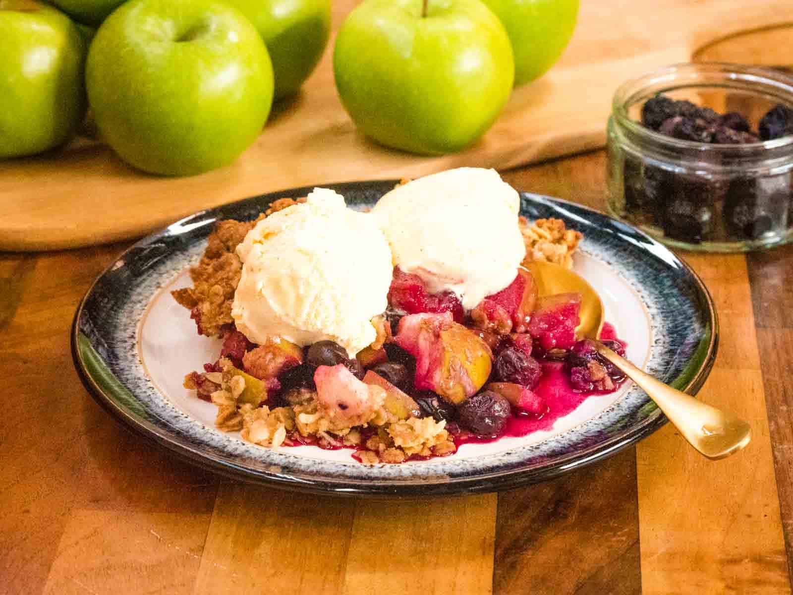 2 scoops of vanilla ice cream on a serving of freshly baked apple berry crumble surrounded by granny smith apples