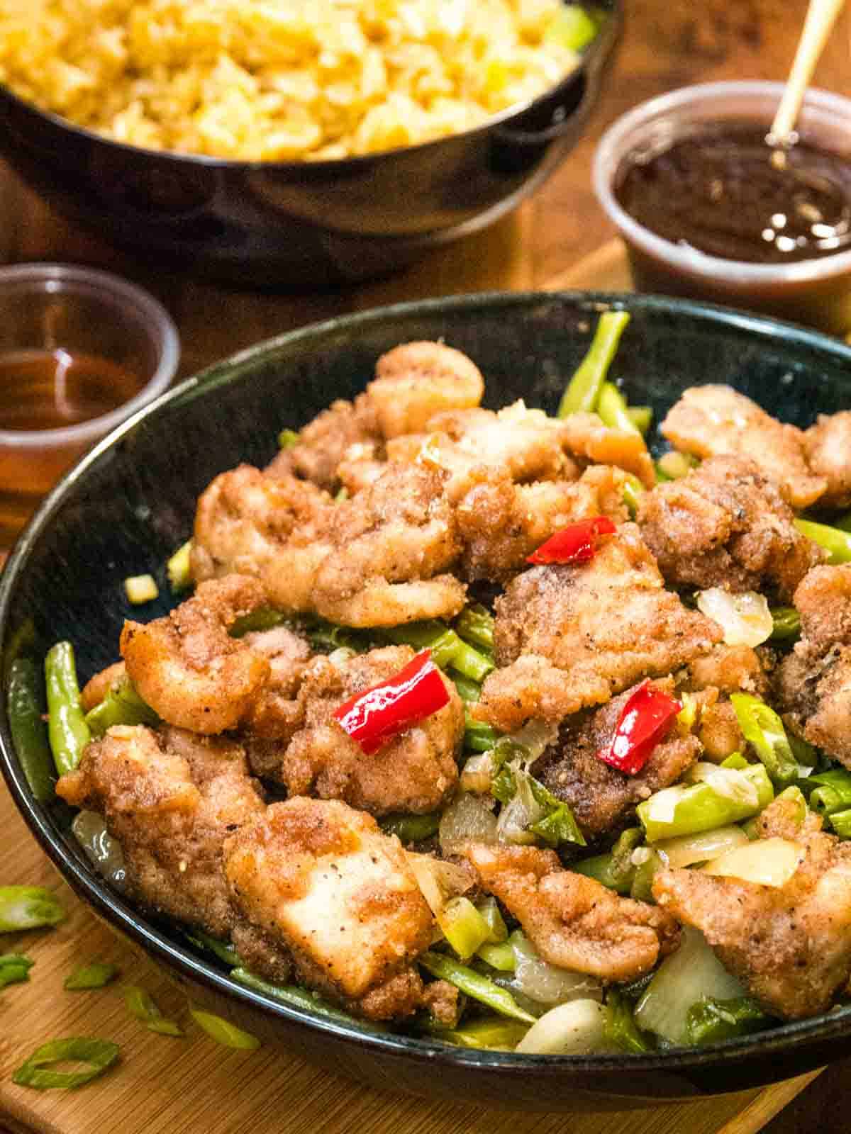 close up of battered salt and pepper chicken with fried vegetables n black bowl with sliced spring onions