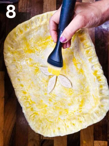 person brushing beaten egg wash on raw puff pastry, including pie leaf decorations