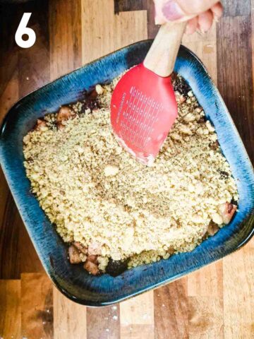 person spreading blackberry crumble with apple cookie crumb topping on top of fruit filling in baking dish with spatula