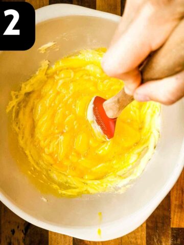 person mixing in eggs into sugar and butter in mixing bowl with wooden spoon