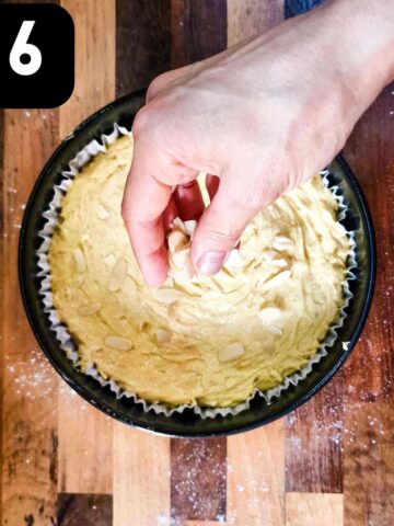 person sprinkling sliced almonds on top of cake in a lined round cake tin
