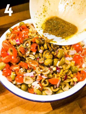 person drizzling a herby vinaigrette over olives, tomatoes, onions and sweetcorn cold pasta underneath in a large serving bowl