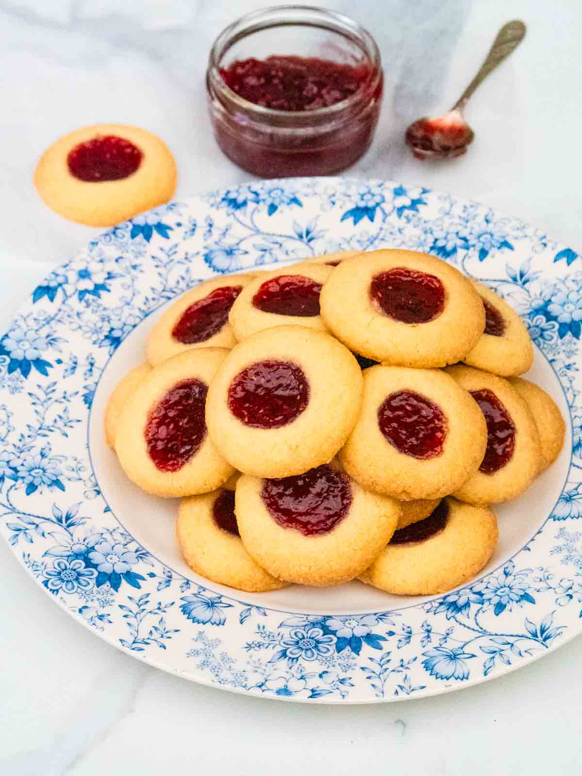 Blue floral serving plate full of jam thumbprint biscuits surrounded by raspberry jam on white marble worktop